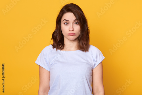 Studi oshot of dark haired young woman dresses casual white t shirt, standing and looking at camera, posing with pouty lips isolated over yellow background, being offended and having bad mood.