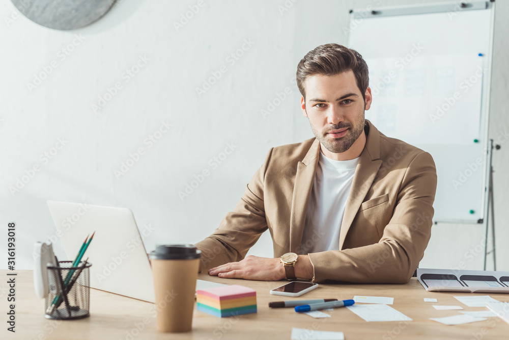 Handsome designer looking at camera while working with laptop and ux website templates in office