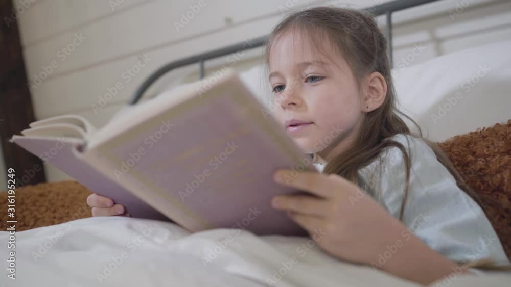 Close-up of little brunette Caucasian girl reading book in bed. Happy charming child enjoying stories at bedtime. Hobby, lifestyle, leisure. Close-up of little brunette Caucasian girl reading book in bed. Happy charming child enjoying stories at bedtime. Hobby, lifestyle, leisure.