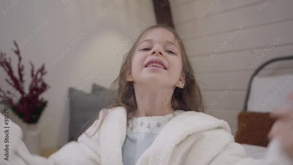 Camera approaching to cute little Caucasian girl in white bathrobe shaking long curly hair. Pretty kid sitting on bed after shower and smiling. Leisure, lifestyle, relaxation. Camera approaching to cute little Caucasian girl in white bathrobe shaking long curly hair. Pretty kid sitting on bed after shower and smiling. Leisure, lifestyle, relaxation.