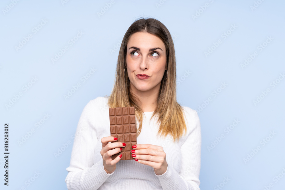 Young blonde woman over isolated blue background taking a chocolate tablet and having doubts Young blonde woman over isolated blue background taking a chocolate tablet and having doubts