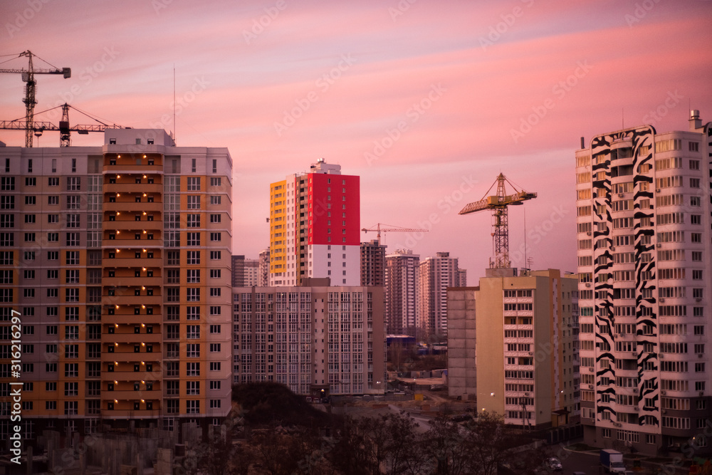 Industrial construction cranes and building silhouettes over sun at sunrise. Unfinished buildings development in pink sunset light. Silhouette of crane in morning light