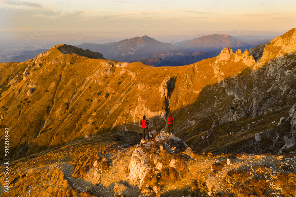 Aerial drone photograph with couple of hikers watching a beautiful sunset on top of Bucegi mountain ridge, during magical golden hour light in autumn season in Romania