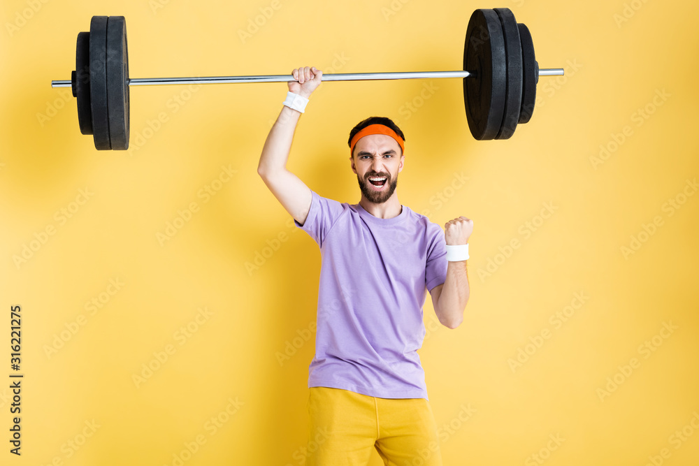 excited sportsman celebrating while working out with barbell on yellow excited sportsman celebrating while working out with barbell on yellow