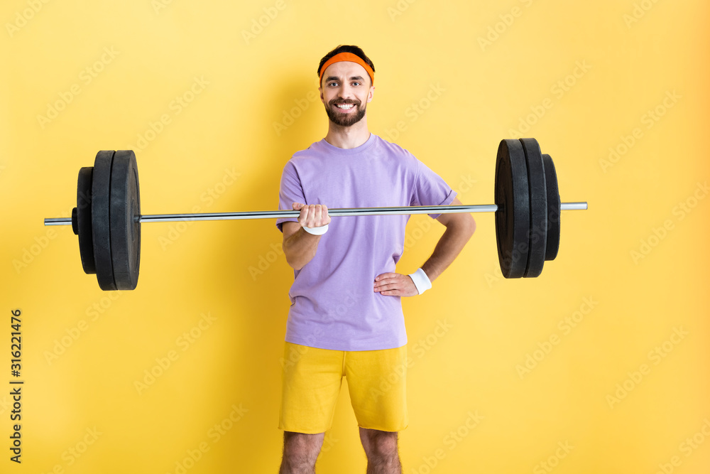cheerful sportsman standing with hand on hip and holding barbell on yellow cheerful sportsman standing with hand on hip and holding barbell on yellow