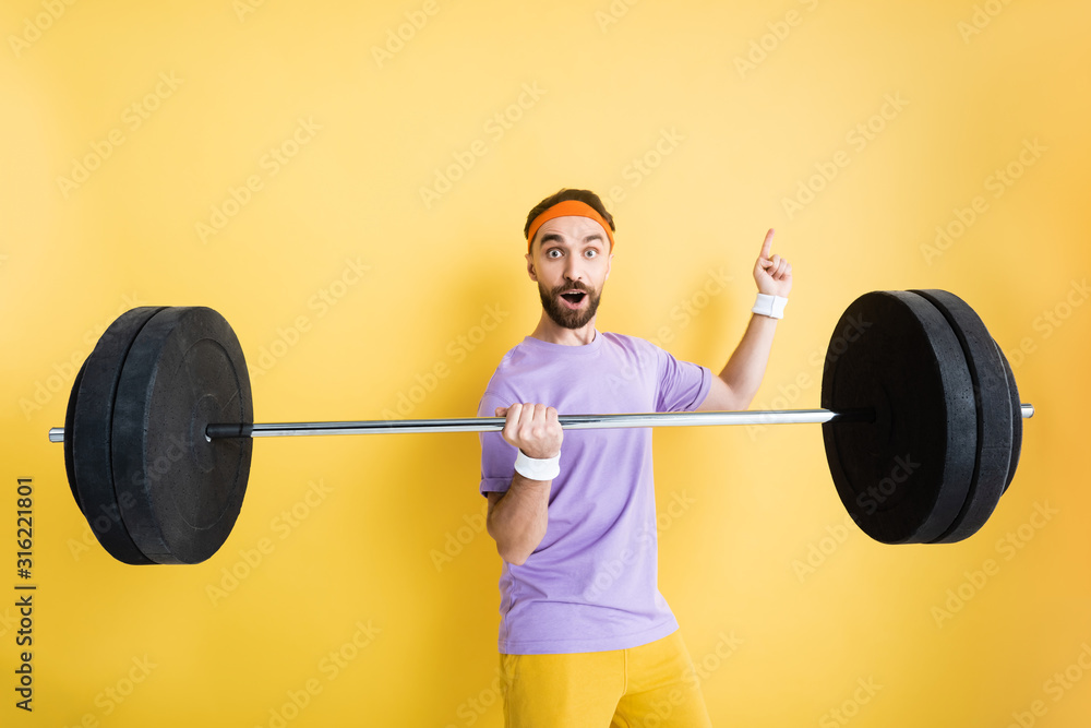 surprised sportsman pointing with finger and holding barbell on yellow surprised sportsman pointing with finger and holding barbell on yellow