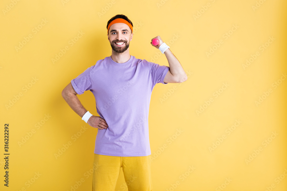 cheerful sportsman holding dumbbell while standing with hand on hip on yellow