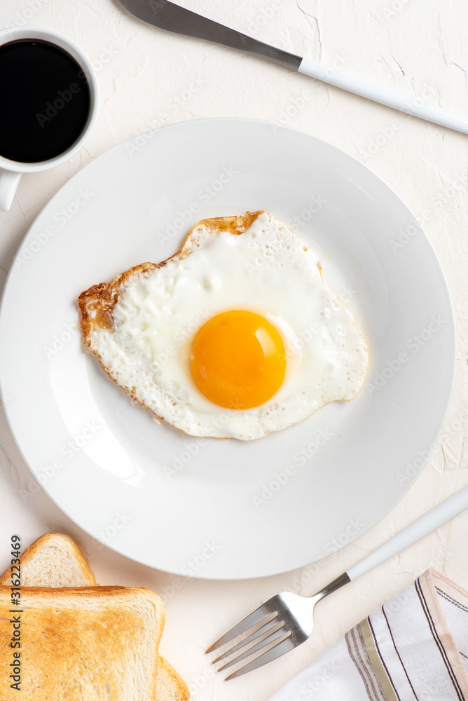 Traditional breakfast. One fried egg on a white plate, toasts and coffee on a white table, top view, flat lay. Traditional breakfast. One fried egg on a white plate, toasts and coffee on a white table, top view, flat lay.