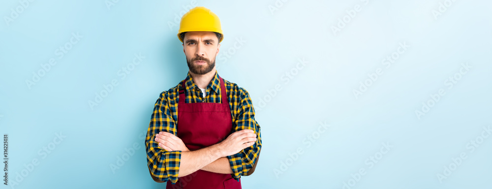 panoramic shot of offended workman in safety helmet standing with crossed arms on blue