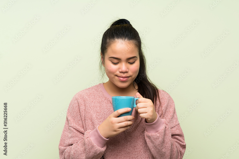 Young teenager Asian girl over isolated green background holding hot cup of coffee Young teenager Asian girl over isolated green background holding hot cup of coffee