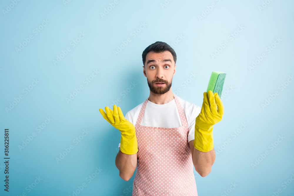 confused bearded man in rubber gloves holding sponge on blue confused bearded man in rubber gloves holding sponge on blue