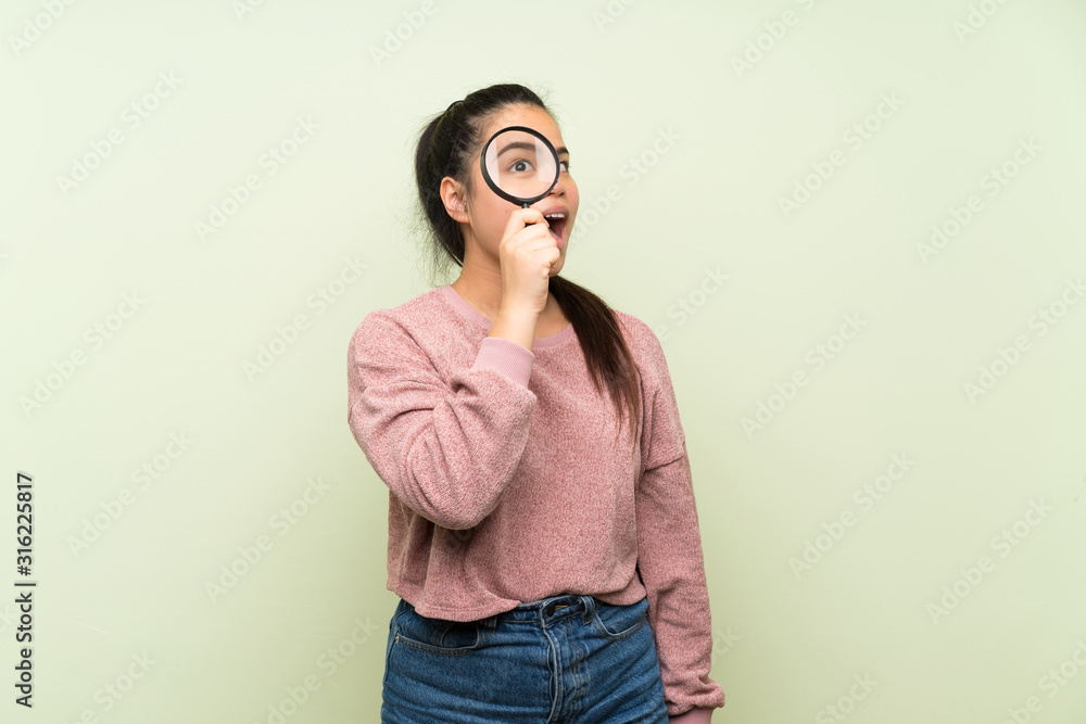 Young teenager Asian girl over isolated green background holding a magnifying glass