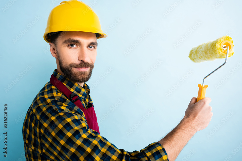 happy handyman in safety helmet holding paint roller on blue