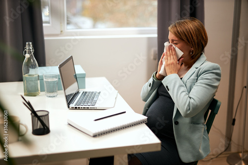 Sick caucasian pregnant businesswoman sitting in her office and blowing her nose.