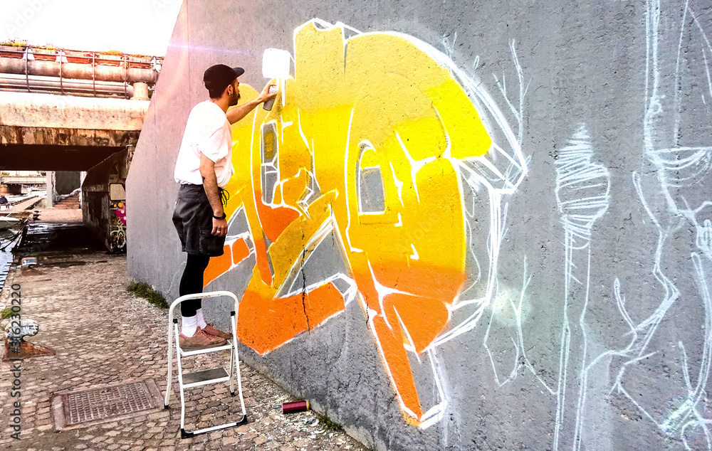 Naklejka premium Artysta uliczny pracujący nad kolorowym graffiti na ścianie w przestrzeni publicznej - Sztuka współczesna przedstawia koncepcję miejskiego faceta malującego na żywo fototapety z żółtym i pomarańczowym aerozolem w sprayu - Jasny filtr odblaskowy