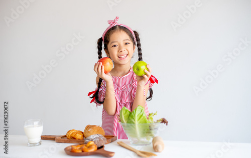 Lovely Asian little girl hold apples and smile with vegetables  milk and bread on table with white wall background.
