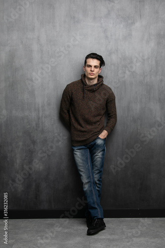 Studio fashion portrait of young attractive man in brown hoodie and blue jeans. Guy stands cross-legged while holding hands in pockets against a gray textured wall with free copy space