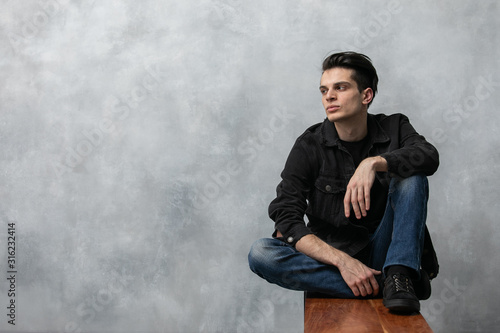 Studio fashion portrait of attractive young man in black jacket and blue jeans sitting on wooden bench against grey textured wall.