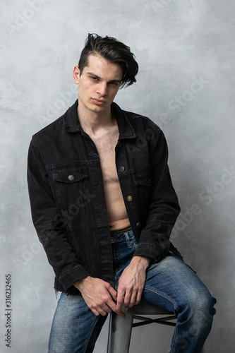 Studio fashion portrait of attractive young man in a black jacket over his naked body. Guy posing against grey textured wall.