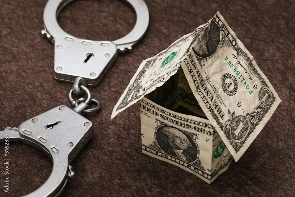 Toy house made of dollars and handcuffs on the table. Real Estate Fraud Concept Toy house made of dollars and handcuffs on the table. Real Estate Fraud Concept