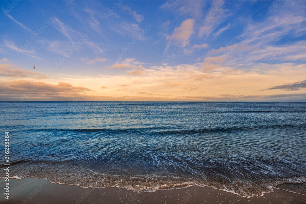 Meer mit Wolken am Horizont in Abendstimmung orange blau und leichte Welle am Strand im Vordergrund