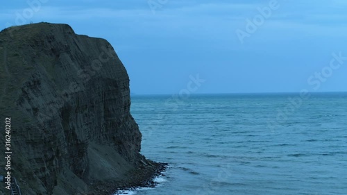 Aerial view of the large grey cliff and sea waves breaking on the rocks against the blue evening sky. Shot. Beautiful evening marine scenery photo