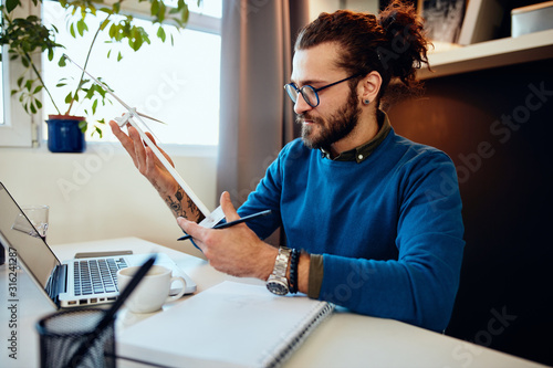 Fotografie Young caucasian bearded engineer sitting in his office, holding windmill model and thinking what how redesign it