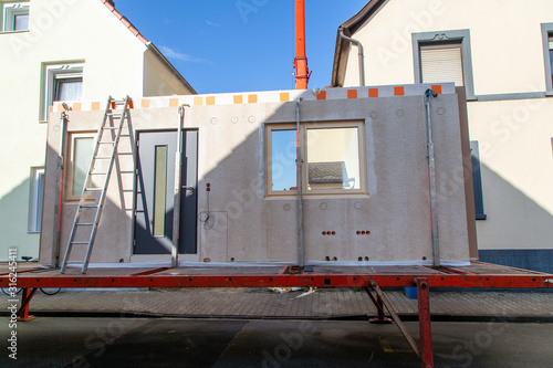 Truck trailer in orange with component for a prefabricated house of a ladder leaning against it and in the background the car crane boom