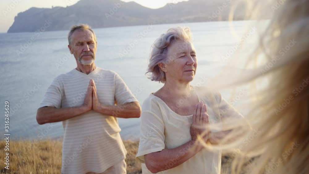 Group of senior people doing yoga exercises at the seaside on the background at the sunset. Feeling the harmony. Concept of calm and meditation. Way to peace and balance. Handheld, close-up shot Group of senior people doing yoga exercises at the seaside on the background at the sunset. Feeling the harmony. Concept of calm and meditation. Way to peace and balance. Handheld, close-up shot