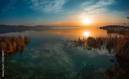 Beautiful sunrise on the lake and mountains. Bright sun rays with fog reflected on the lake with the reeds on the shore.
