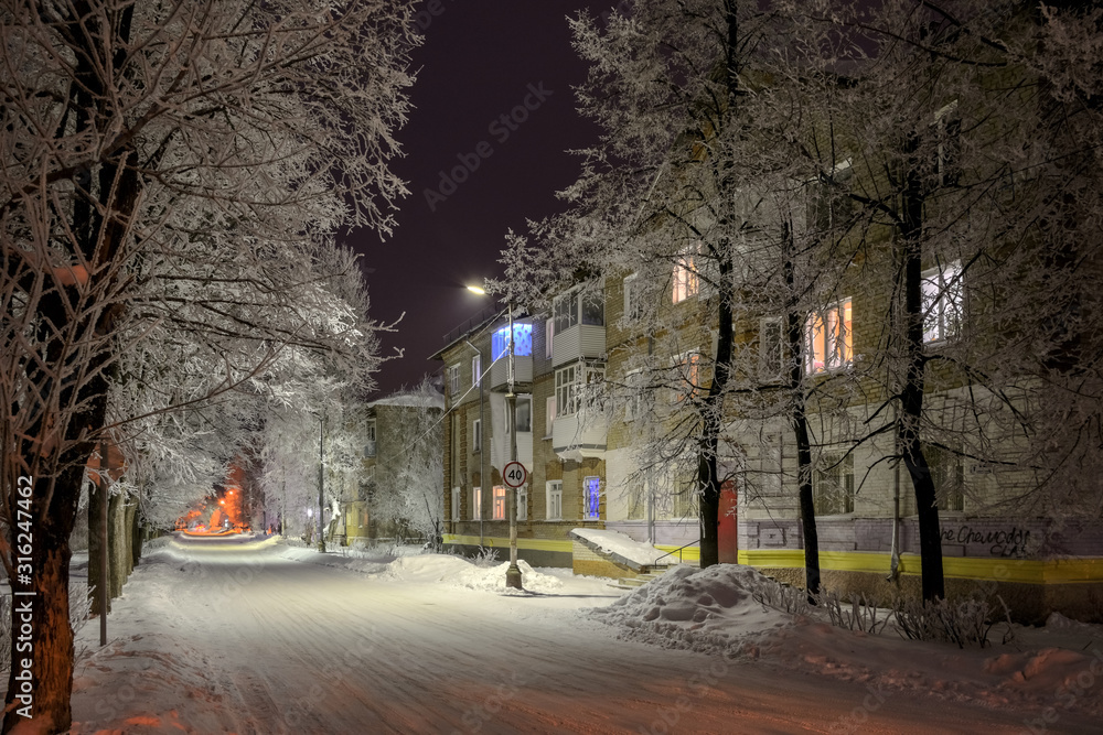 Night street with snowy trees in the light of lanterns and windows of houses, painted in different colors.