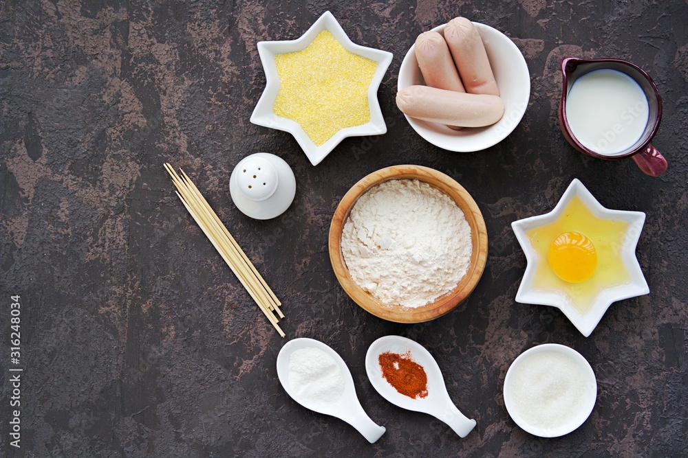 Ingredients for making corn dogs on a dark concrete background : sausages, corn flour, wheat flour, egg, sugar, baking powder, salt, wooden skewers, paprika, milk. Ingredients for making corn dogs on a dark concrete background : sausages, corn flour, wheat flour, egg, sugar, baking powder, salt, wooden skewers, paprika, milk.