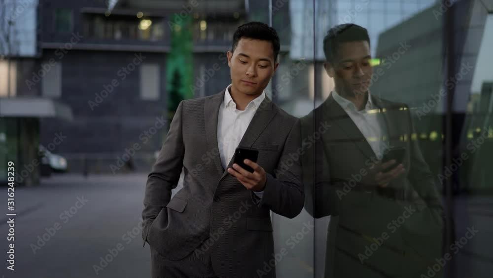 Real time portrait shot of a young asian businessman conducting business correspondence, standing leaning against the wall. Real time portrait shot of a young asian businessman conducting business correspondence, standing leaning against the wall.