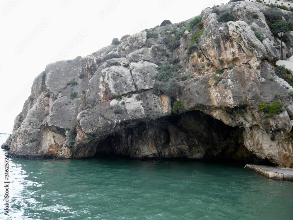Rock clinging shrubbery on overhanging cliff of eroding shallow cave in turquoise water . 