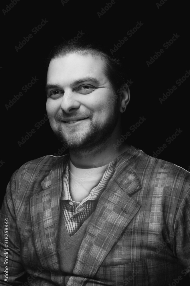 black and white low-key portrait of cheerful handsome bearded stylish elegant man in plaid blazer smiling and looking away on dark background