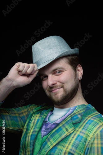 low-key portrait of handsome bearded smiling man in green checkered blazer putting on stylish grey hat isolated on black  © Viktoriia