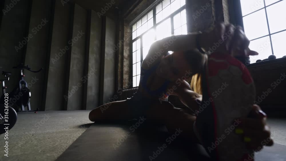 Young athletic woman warming up before workout in the gym. Lateral motion shot, silhouette girl stretching her legs in backlit. Flexibility girl doing exercising on floor of modern loft. Bottom view Young athletic woman warming up before workout in the gym. Lateral motion shot, silhouette girl stretching her legs in backlit. Flexibility girl doing exercising on floor of modern loft. Bottom view