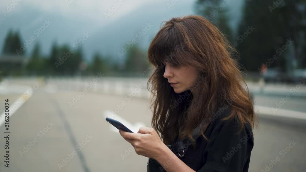 Gimbal shot of woman typing with the phone typing texting standing on blurred background of highway road car lanes and mountains with forest Gimbal shot of woman typing with the phone typing texting standing on blurred background of highway road car lanes and mountains with forest