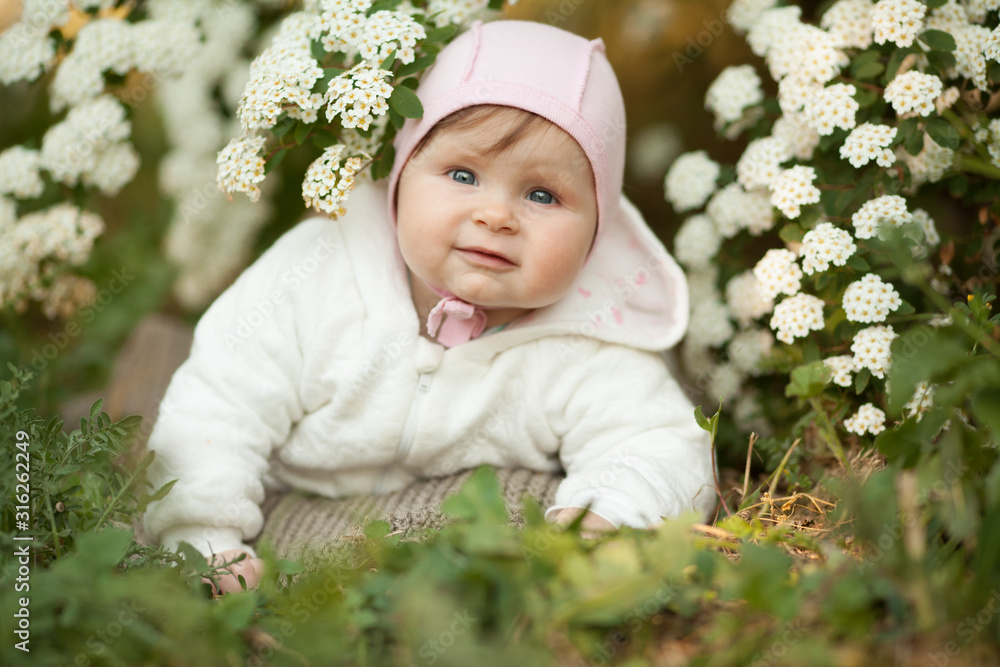 Cute little baby girl in spring in a blooming garden smiling, looking at the camera. Concept: spring, family relations, children. The care for the baby. Cute little baby girl in spring in a blooming garden smiling, looking at the camera. Concept: spring, family relations, children. The care for the baby.