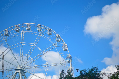 white ferris wheel carousel on a background of blue blue sky with white clouds and green foliage on trees summer