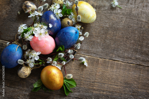 Colored Easter eggs in a nest with willow branches and spring flowers on a gray wooden background. Top view flat lay background. Copy space.