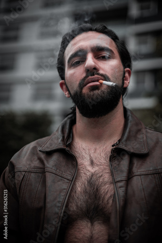 Well-built latino model, handsome brunet men with beard, dark eyes and cigarette in unbuttons a leather jacket shows his chest poses and smokes in the city center of Buenos Aires. Street portrait.