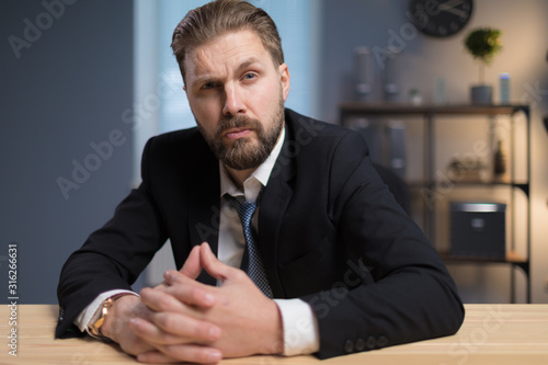 Serious bearded businessman sitting at desk, his fingers interlocked and eyebrow raised