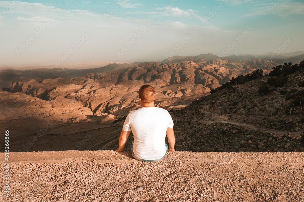 A lone man sitting on a rock and watching something on the horizon of an island in the Norwegian fjords.He's wearing a white t-shirt.The sun shines on the canyon. thoughts of infinity.sunny weather