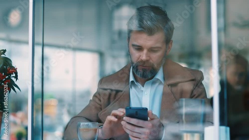 View through the window of attractive serious middle aged businessman using phone at coffee shop texing message working watching video successful people daily routine entertaining communicating