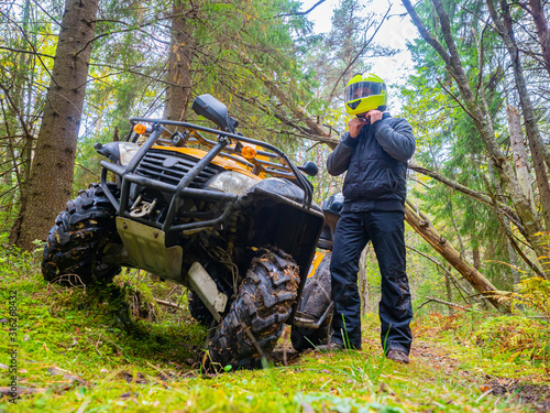 A man in a yellow helmet stands next to ATV. ATV riding in the woods. Off-road transport. The equipment of the driver of the ATV. Racing on the forest road.