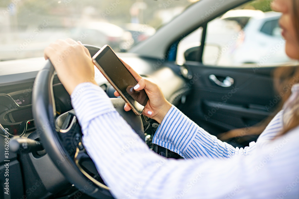 Close up of female hand holding smatrphone and typing while driving. Woman texting message using cellphone sitting in car. Answering call while being at wheel. Hand with phone on car cabin background