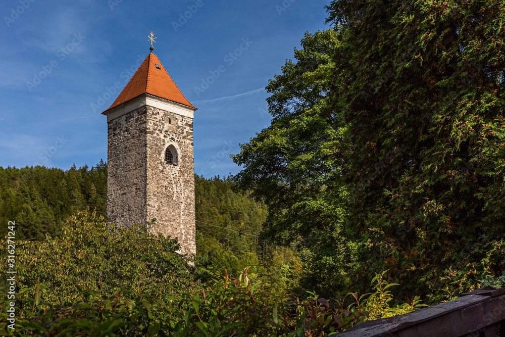 Nejdek / Czech Republic - September 15 2019: A stone tower with red roof and golden cross is a part of former castle built in 14th century. It is surrounded with green trees. Sunny day with blue sky.
