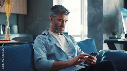 Gorgeous middle aged man with grey hair sitting on the sofa using smartphone in the modern apartment texting message scrolling tapping technology isolated lifestyle slow motion