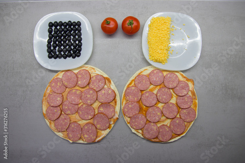 two round pizzas with salami cheese and olives tomatoes and sauce and on a gray table with texture top view
